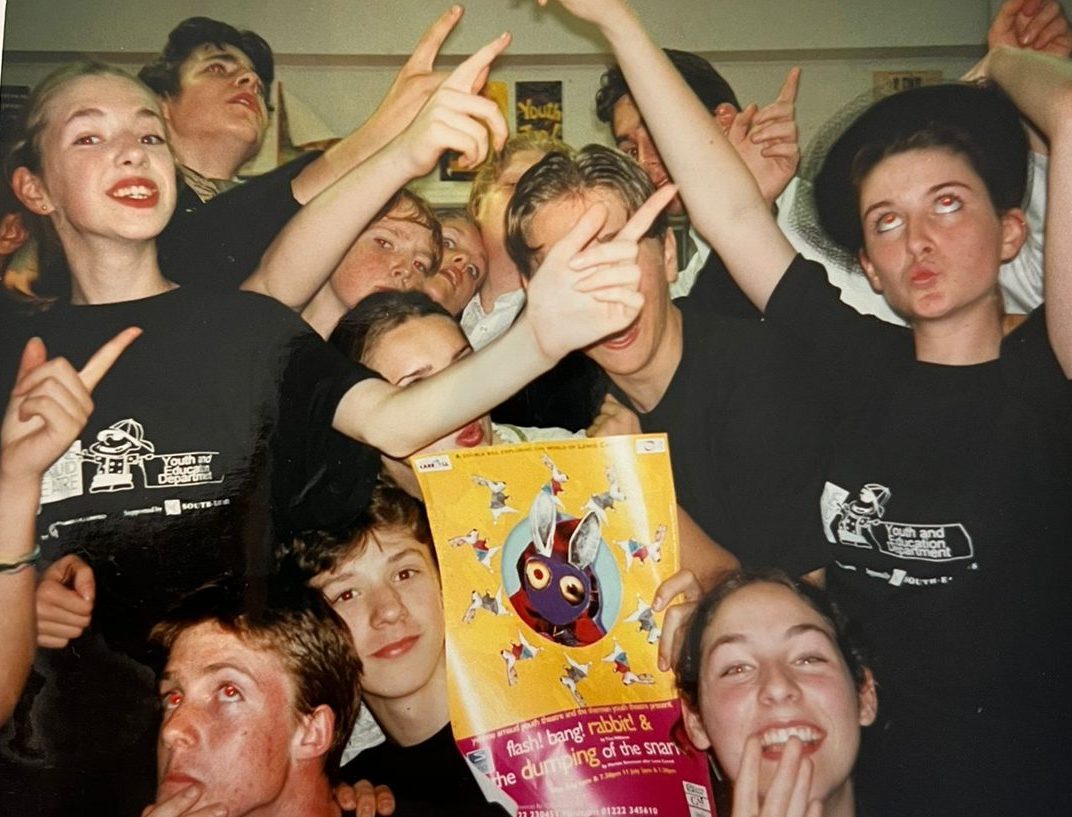 Youth theatre members in black company T-shirts crowding into the frame, pulling faces and holding up a show poster