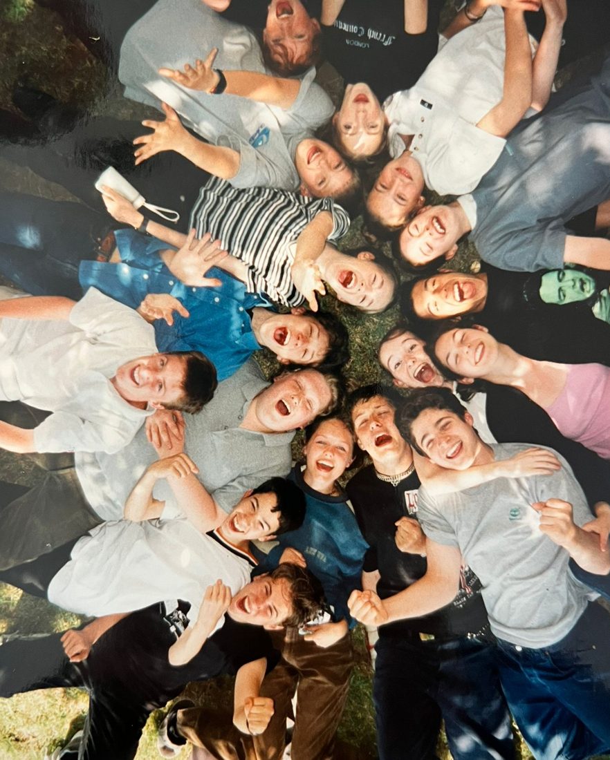 Youth theatre members lying in a circle on the grass, heads together, laughing up at the camera