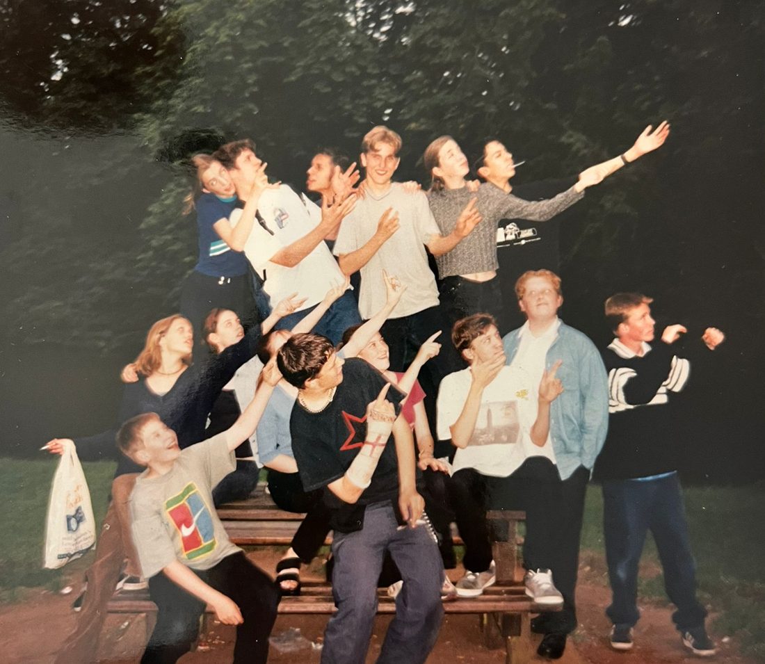 A group of youth theatre members on a park bench at dusk, all striking the same dramatic pointing pose